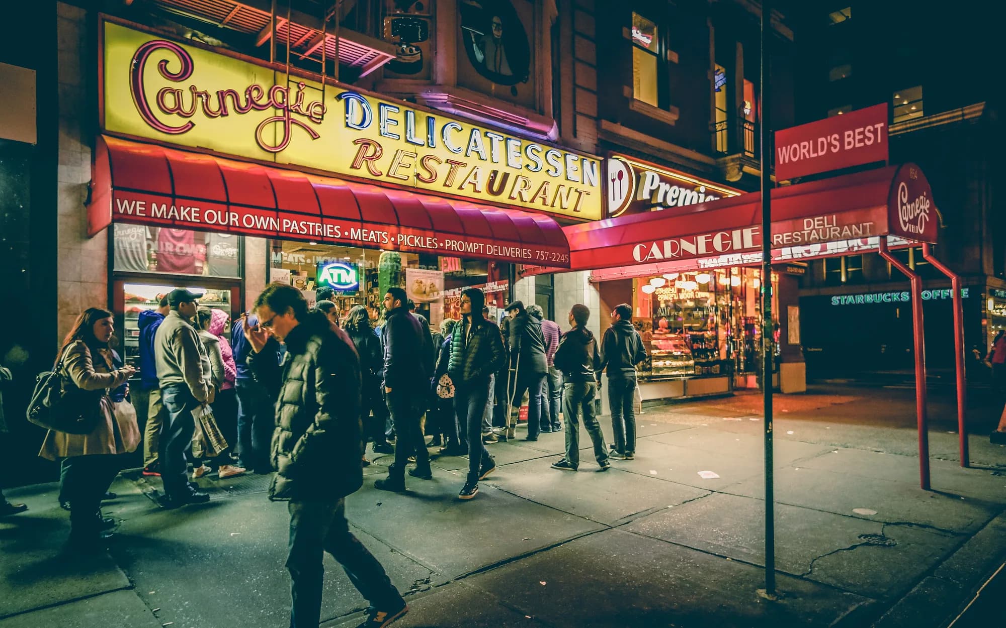 A busy New York City street at night with a lit deli storefront and people passing by